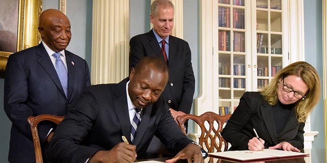 Minister Konneh and MCC chief executive officer Dana J. Hyde sign the deal while Vice President Boakai and a US official look on